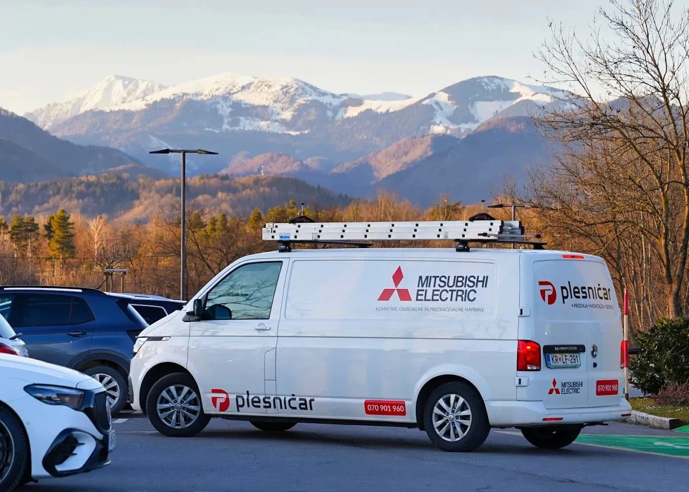Plesničarjev kombi z napisoma Plesničar in Mitsubishi Electric na parkirišču, v ozadju zasnežene gore